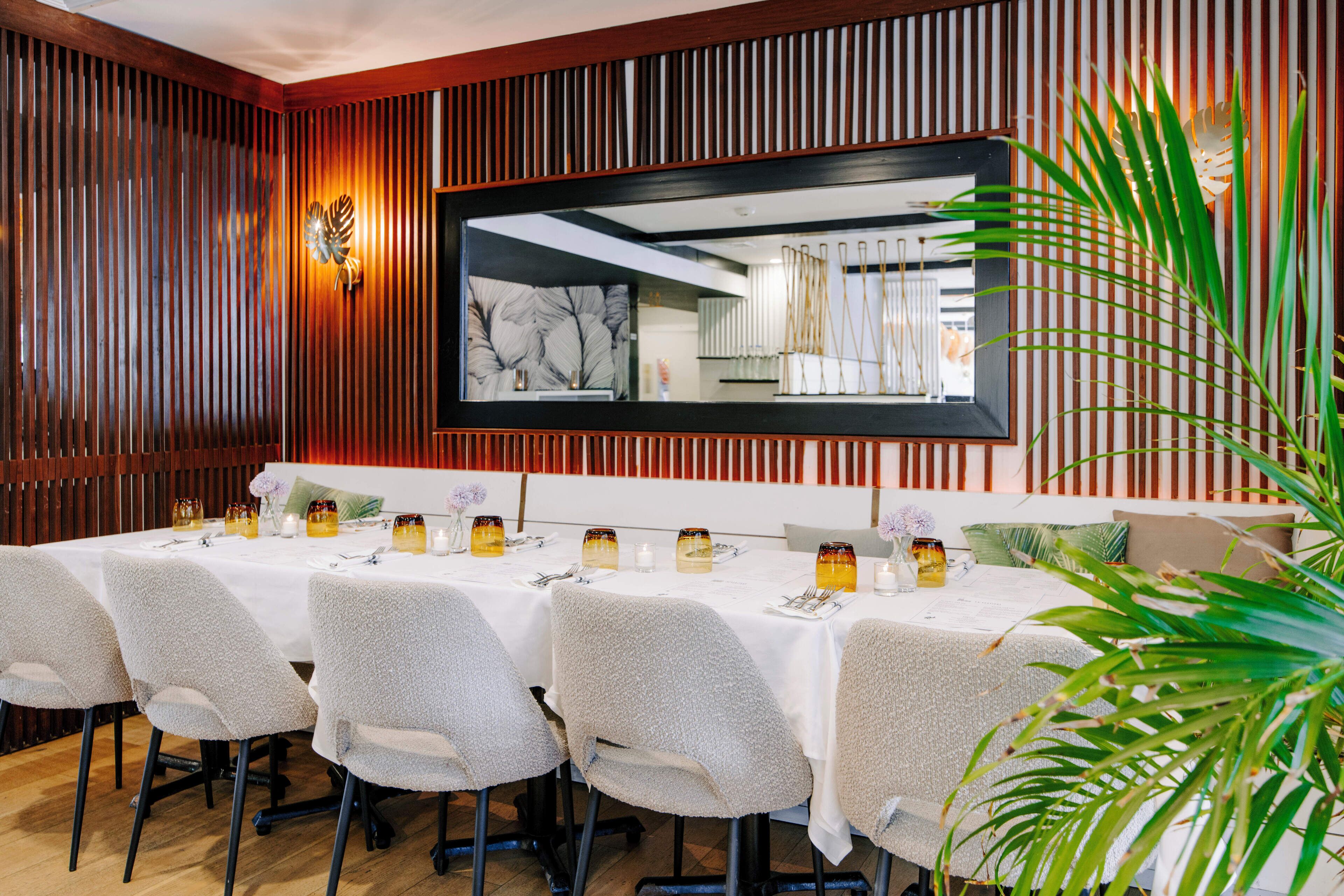A dining area with a long table set for six, surrounded by chairs, and a large mirror on the wall.