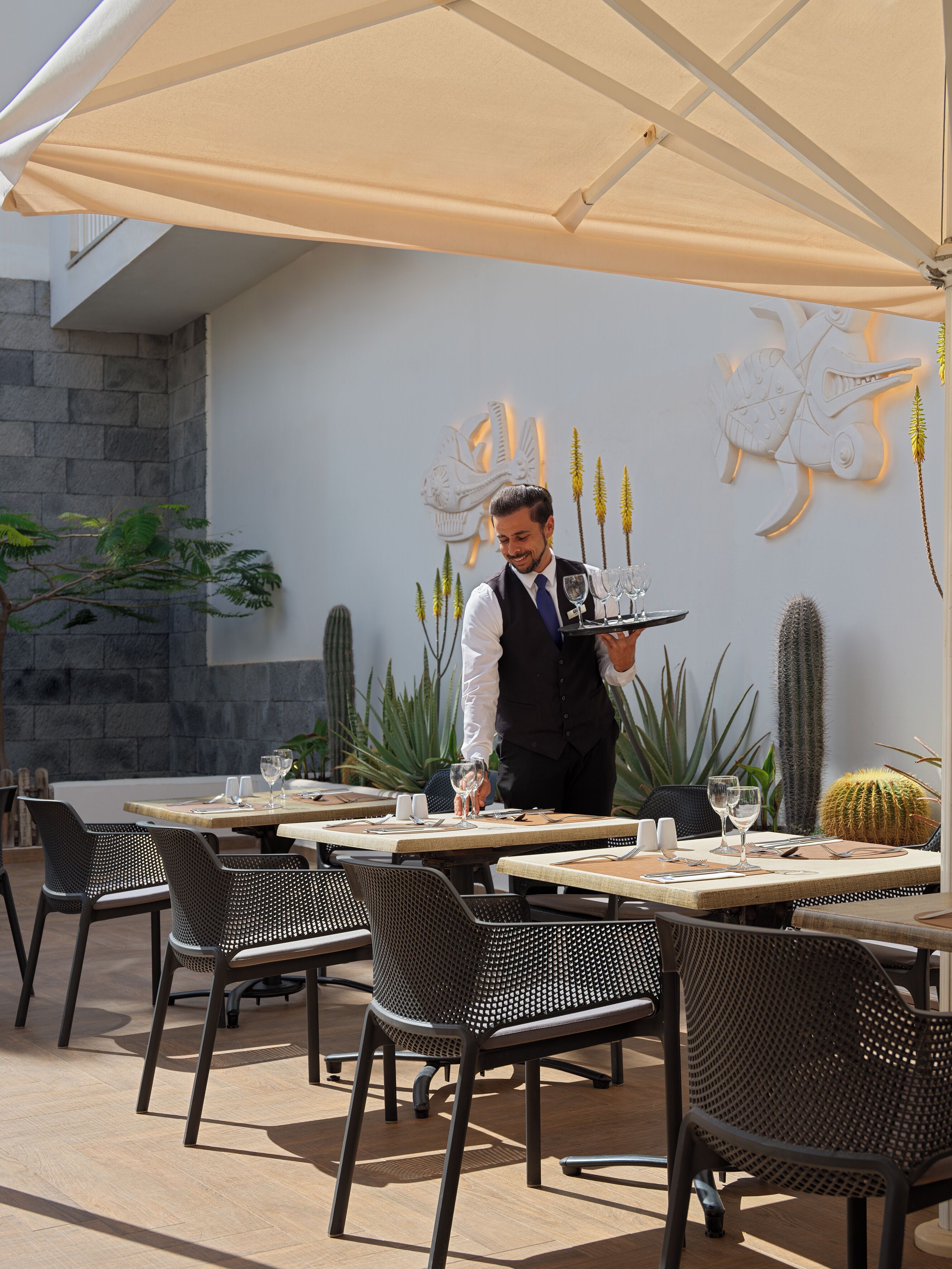 An outdoor dining area with tables and chairs, a waiter serving, and decorative wall art.