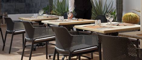 An outdoor dining area with tables and chairs, a waiter serving, and decorative wall art.