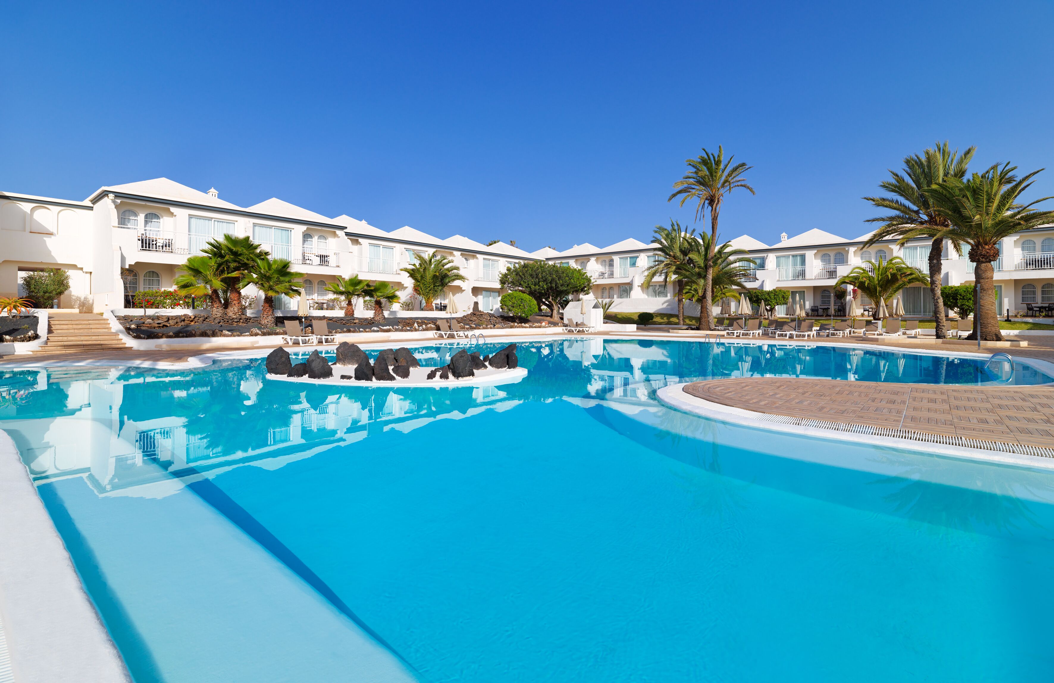 A large swimming pool with lounge chairs and palm trees, surrounded by multi-story hotel buildings.
