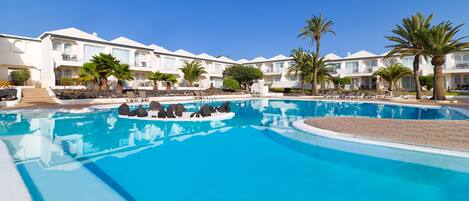 A large swimming pool with lounge chairs and palm trees, surrounded by multi-story hotel buildings.