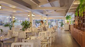A restaurant with white chairs, tables set with wine glasses, and a bar area with wooden cabinets.