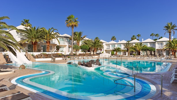 Ett poolområde på ett semesterhotell med flera pooler, palmer och en vit byggnad med balkonger.