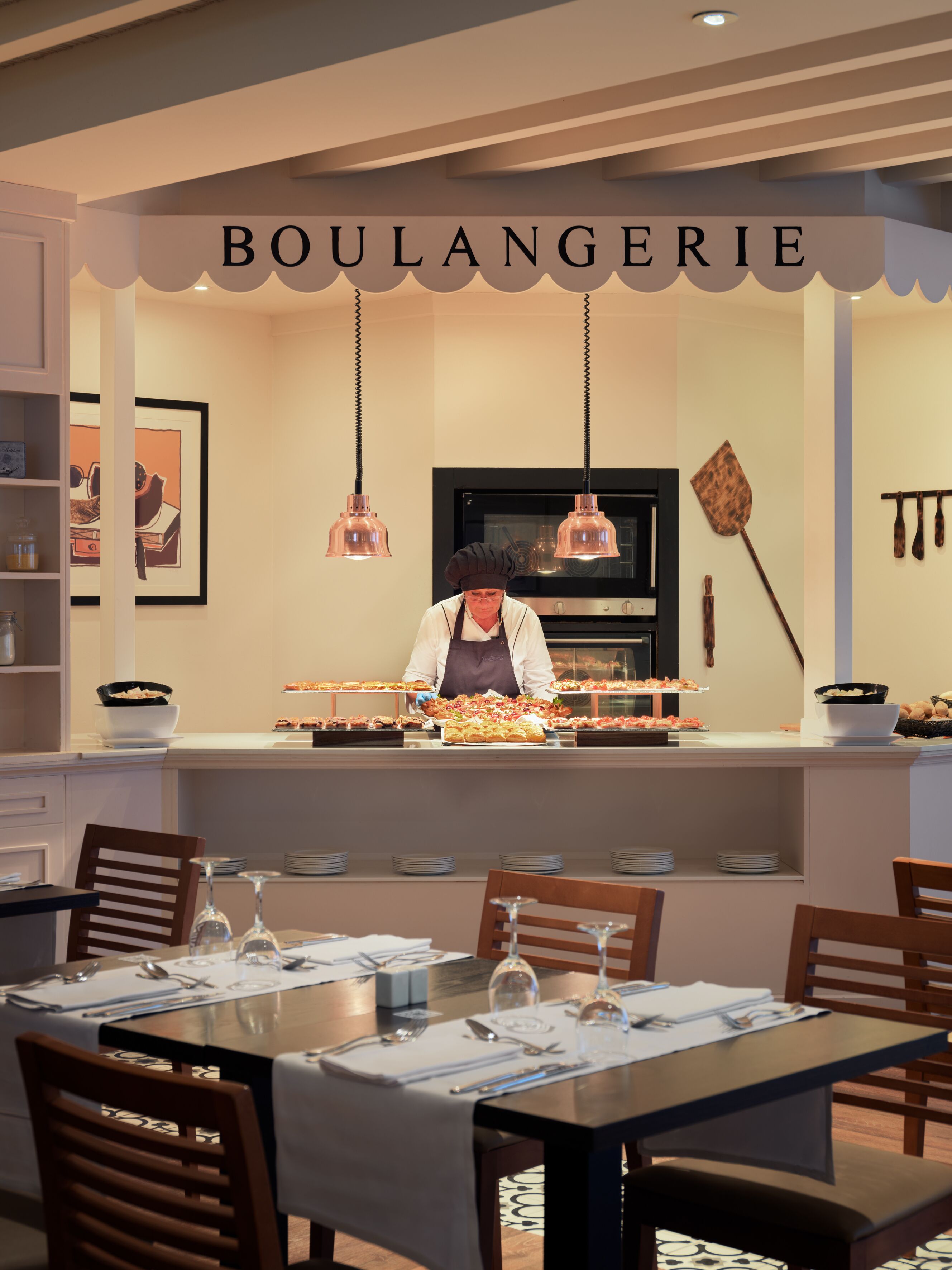Un restaurante con un chef preparando comida en la barra, un bufé con diversos platos, y un comedor con mesas dispuestas para los comensales.