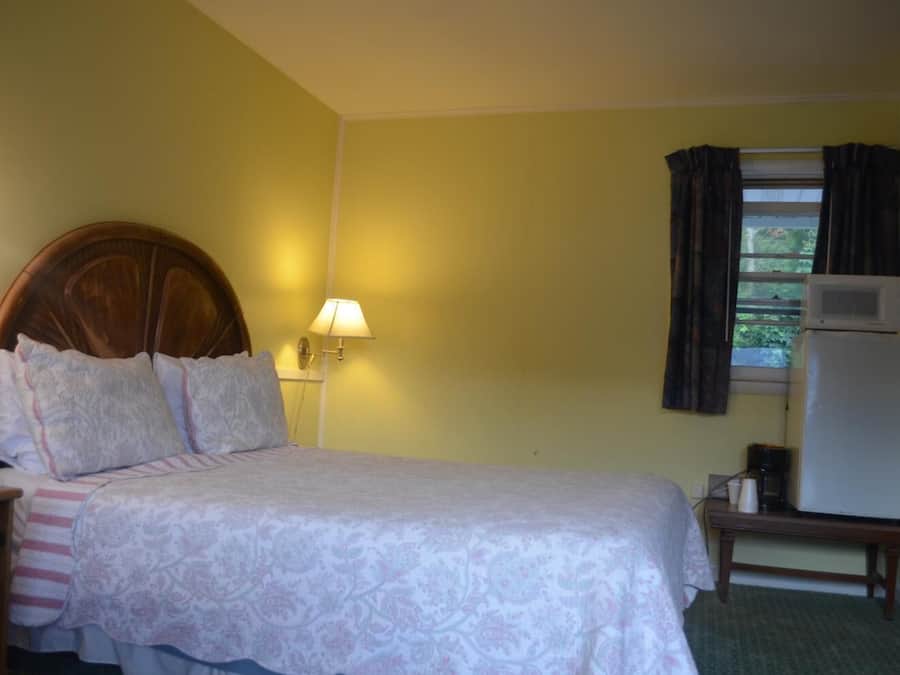 A hotel room with a bed, a nightstand, a lamp, a window with curtains, and a refrigerator.