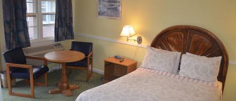 A hotel room with a bed, a desk with a chair, a lamp, and a window with curtains.