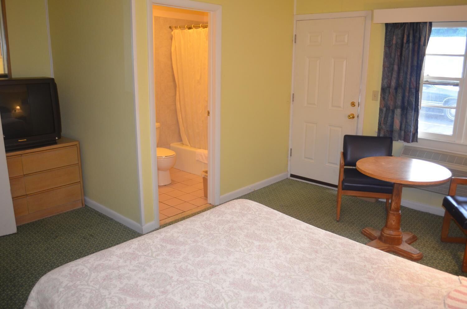 Une chambre d’hôtel comprenant un lit, une petite table, une chaise, un téléviseur, une salle de bain avec un rideau de douche et une porte.
