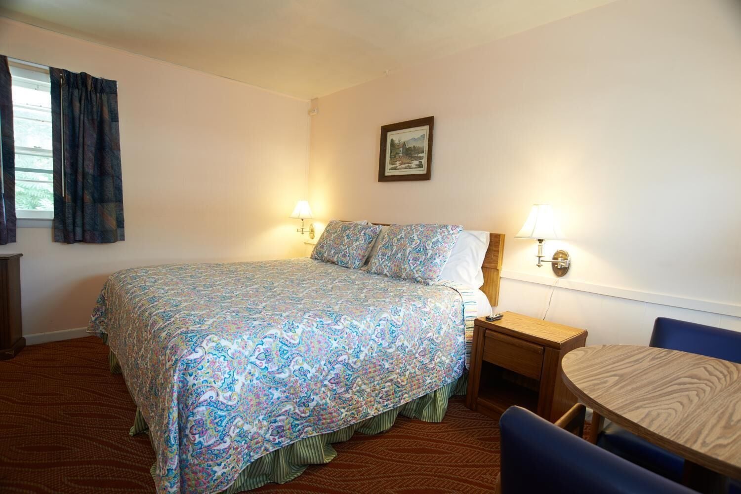 Habitación de hotel con cama, lámparas de noche, un cuadro en la pared, una mesita y una silla.