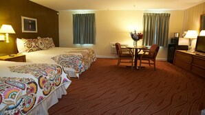 Habitación de hotel con una cama grande, dos mesitas de noche, un escritorio con sillas, un televisor y una pequeña mesa con flores.