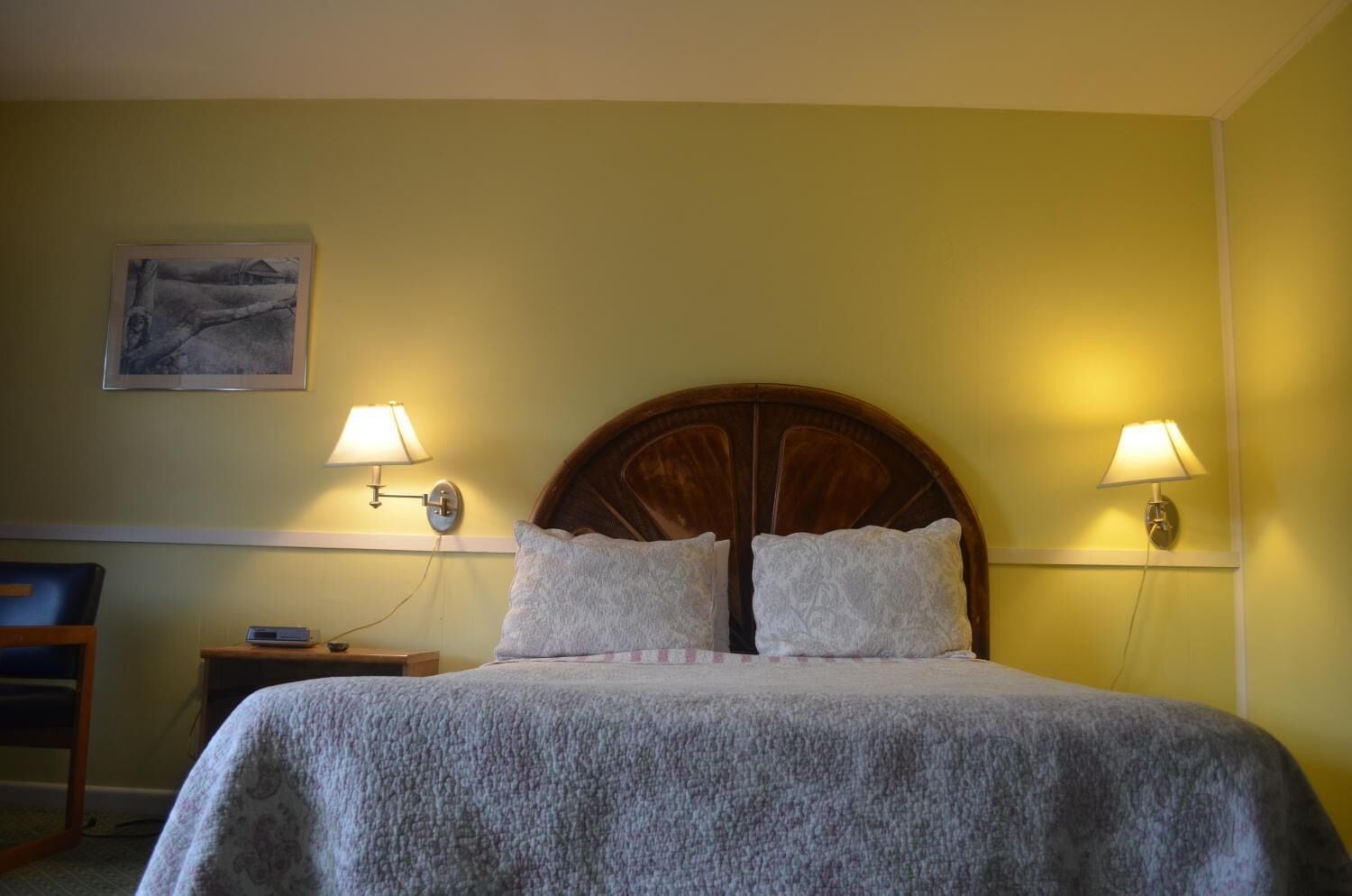 A neatly made bed with a wooden headboard, two bedside lamps, a small bedside table with a lamp, and a framed picture on the wall.