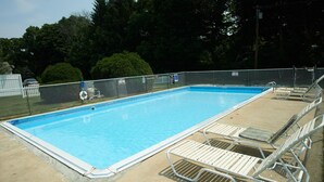 Una piscina rectangular con área cercada, rodeada de árboles y un deck de concreto con tumbonas de metal blanco.