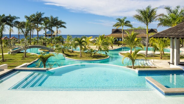 Zona de piscina en un resort con varias piscinas, palmeras y vistas a la playa.