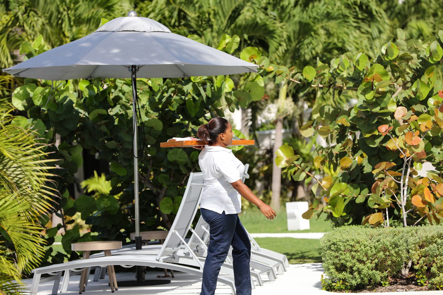 A person carrying a tray with a drink, standing near a white outdoor seating area with a large umbrella.