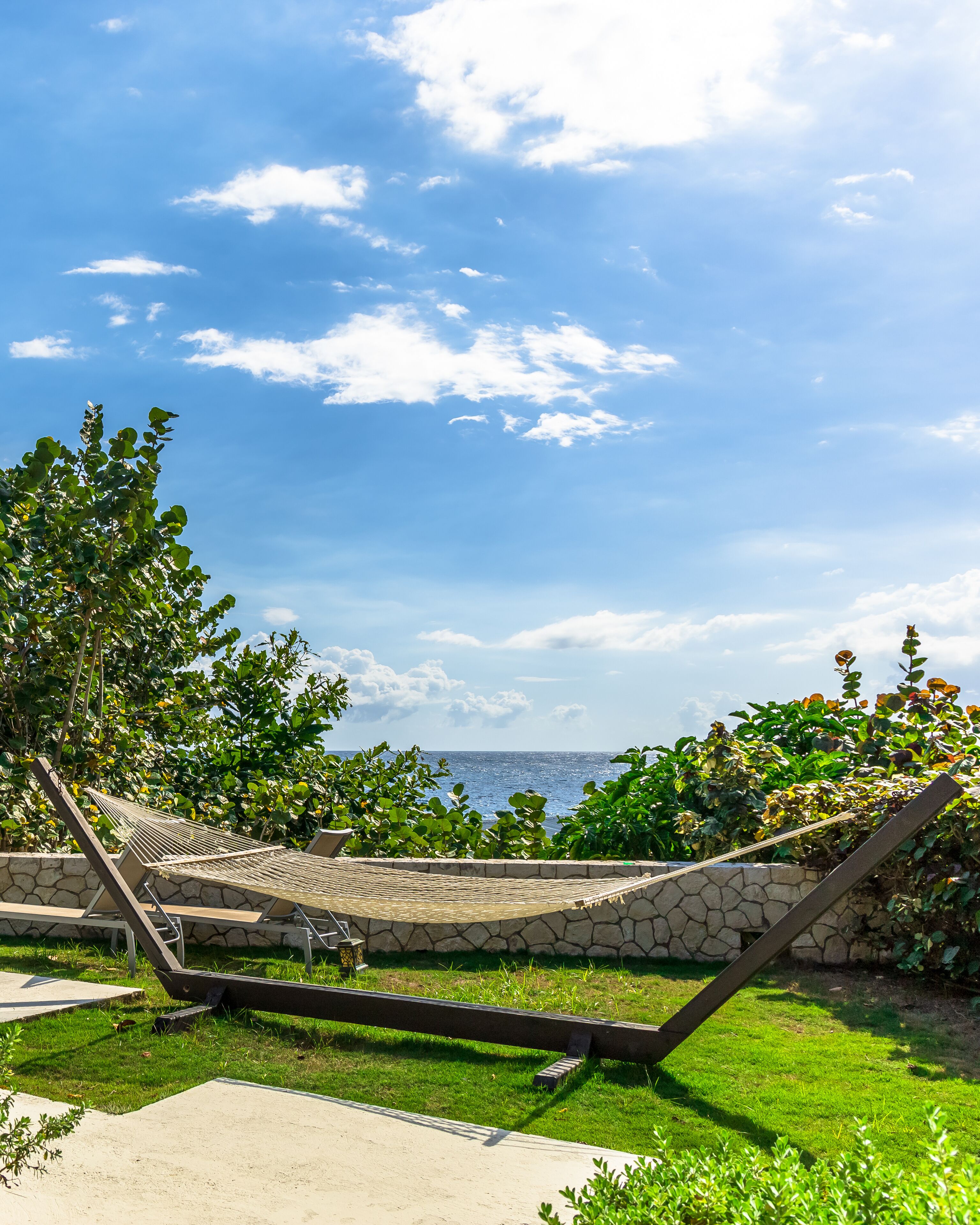 A hammock in a grassy area with a stone wall and plants, overlooking a body of water.