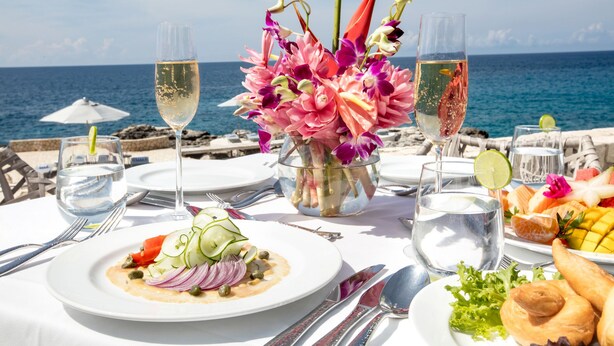 A table set for a meal with a view of the sea, including plates of food, drinks, and a floral centerpiece. - The Cliff Hotel (Negril)