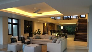 Un salón amplio con un sofá modular grande, dos otomanas y un ventilador de techo. Se ve una escalera que lleva a un nivel superior con un balcón.