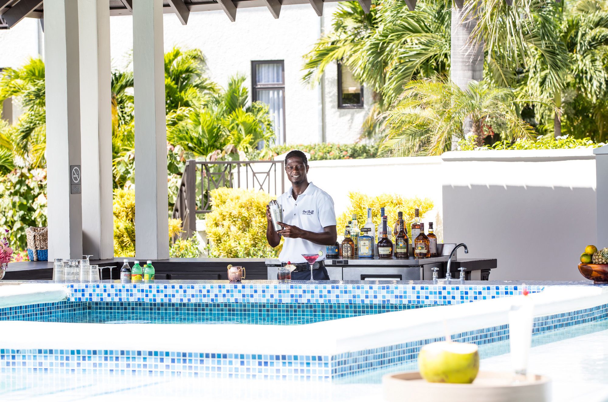 A poolside bar with a bartender, a tiled pool, and tropical plants in the background.