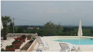 Une piscine entourée de plantes en pot, avec des chaises longues blanches et un parasol blanc, offrant une vue imprenable sur un paysage magnifique.