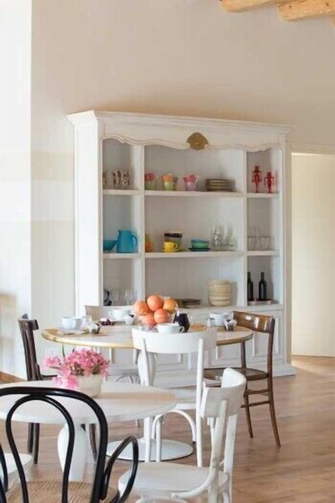 Un coin repas avec un buffet blanc rempli d’assiettes et de verres, une table ronde entourée de chaises et un vase avec des fleurs.