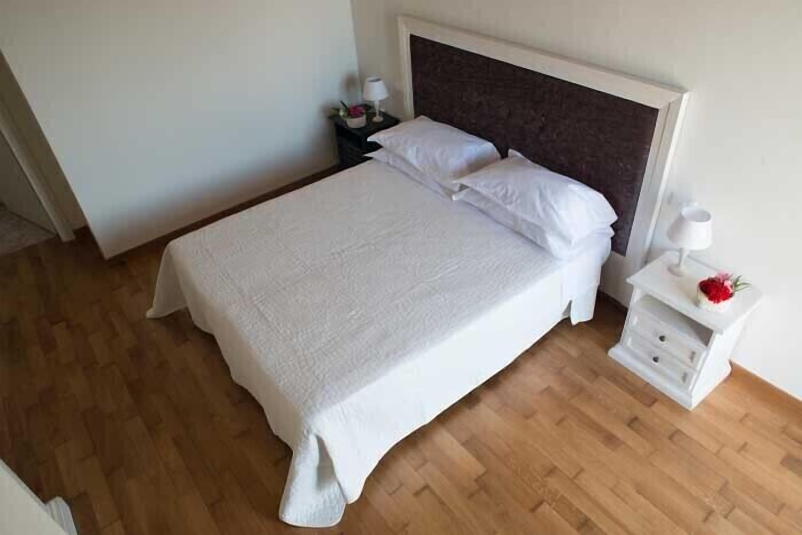Una cama bien hecha con ropa blanca, piso de madera y una mesita de noche blanca con una lámpara y un pequeño objeto decorativo.