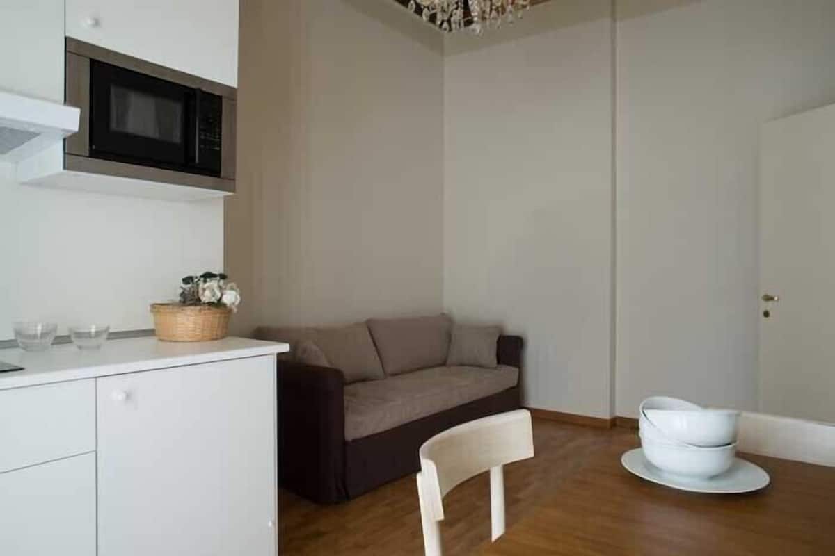 Un espacio habitable compacto con kitchenette, un sofá, una mesa de comedor con una silla y una puerta blanca.