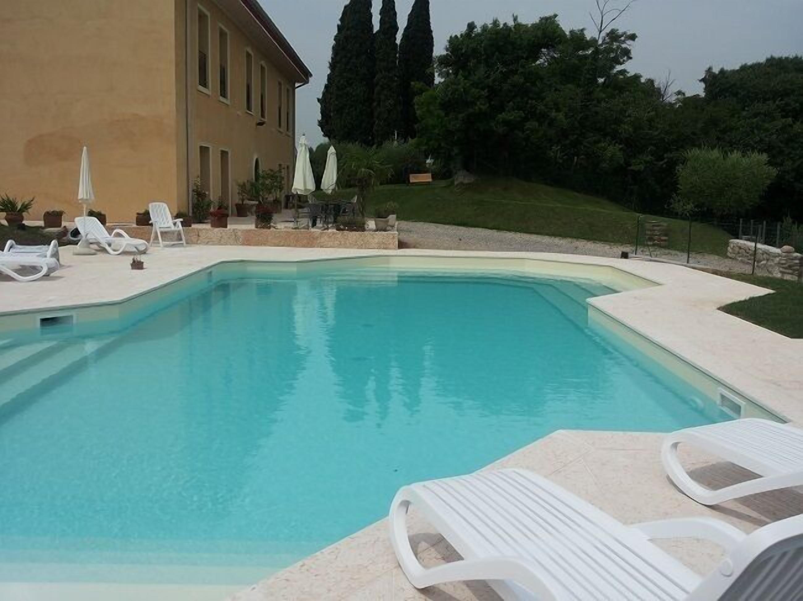 A swimming pool with lounge chairs and umbrellas, surrounded by a building and trees.