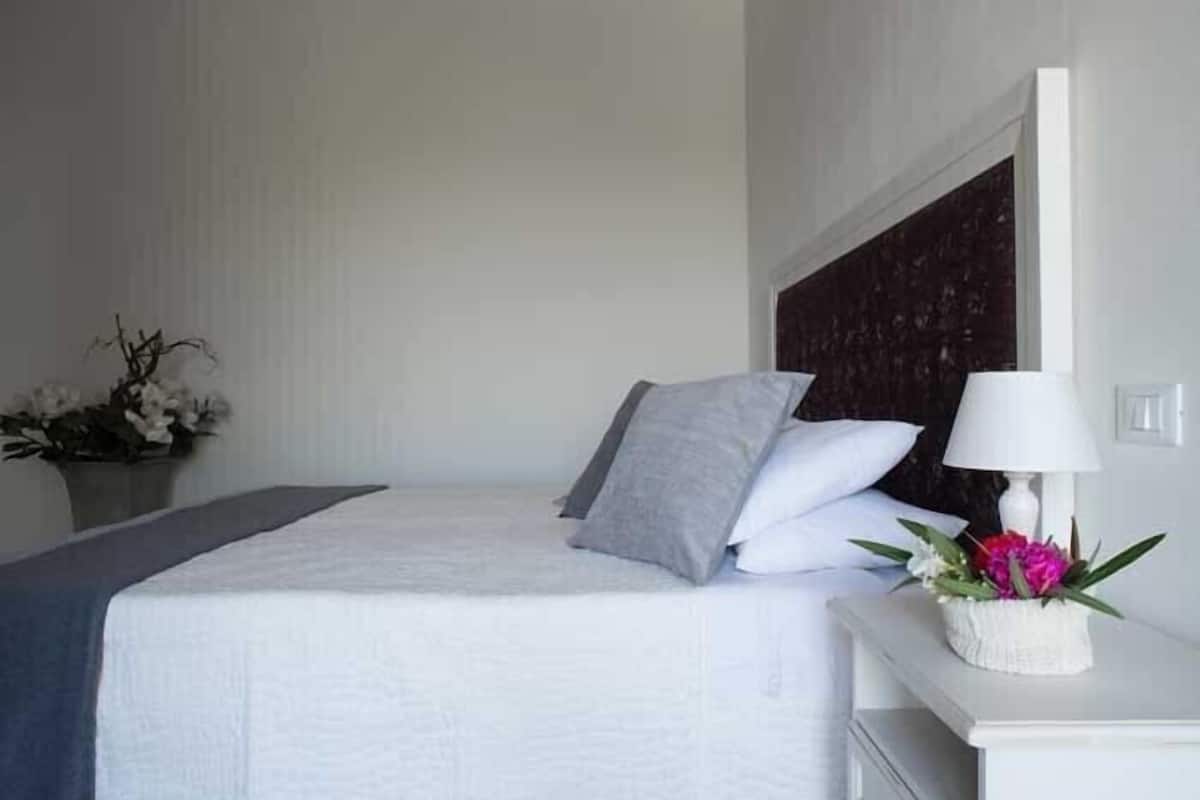 Una cama bien hecha con ropa de cama blanca y gris, una mesita de noche con una lámpara y un arreglo floral, y un cabecero fijado a la pared.