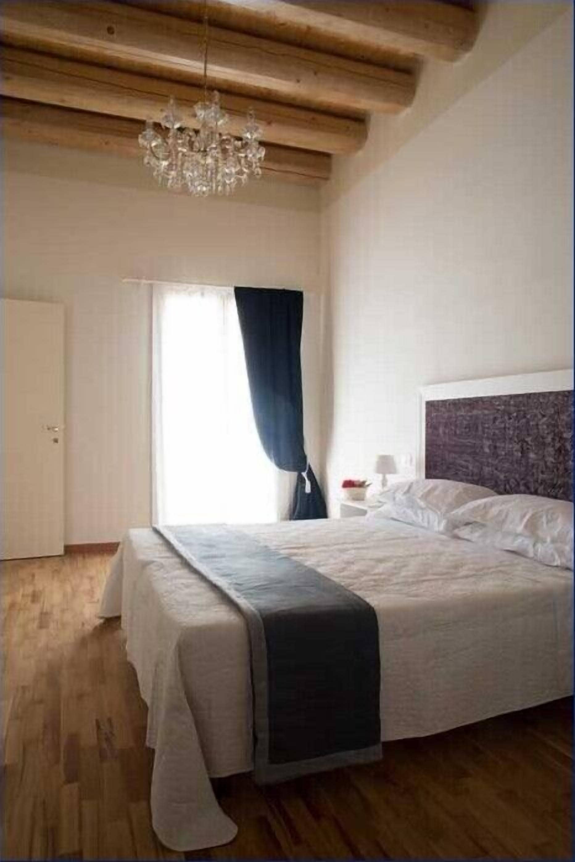 Un dormitorio con una cama grande, vigas de madera en el techo, un candelabro, una ventana con cortinas y una mesita de noche con lámpara.