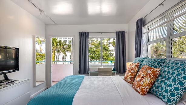 A modern hotel room with a large bed, a TV, and a balcony with a view of palm trees and buildings. - Nobleton Hotel (Fort Lauderdale)