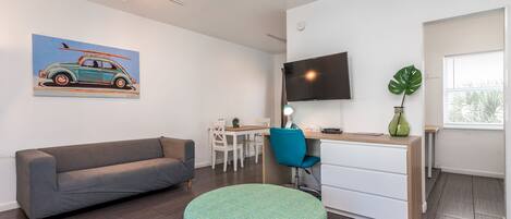 A modern living room with a grey sofa, a round green ottoman, a dining area with a table and chairs, a TV mounted on the wall, and a large plant.
