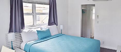 A hotel room with a bed, a window with curtains, a bedside table, and a wall-mounted air conditioning unit.
