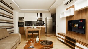 A modern kitchen with a dining area, a TV mounted on the wall, and a unique guitar-shaped coffee table. - Lara Orkide Homes (Antalya)