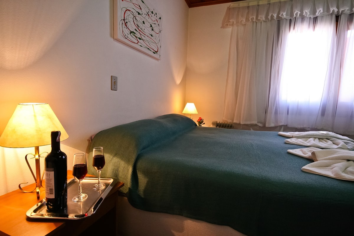 A neatly made bed with a green bedspread, a tray with a bottle of wine and two glasses, and a lamp on a bedside table.