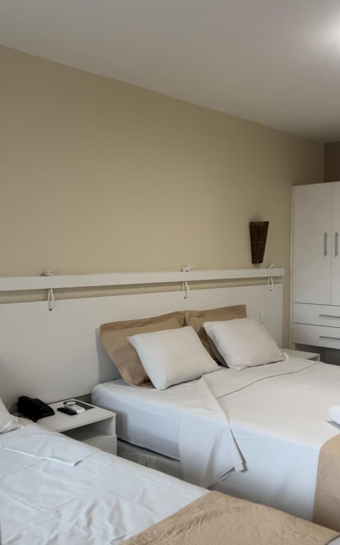 A hotel room with two beds, a wardrobe, and a bedside table with a phone and charger.