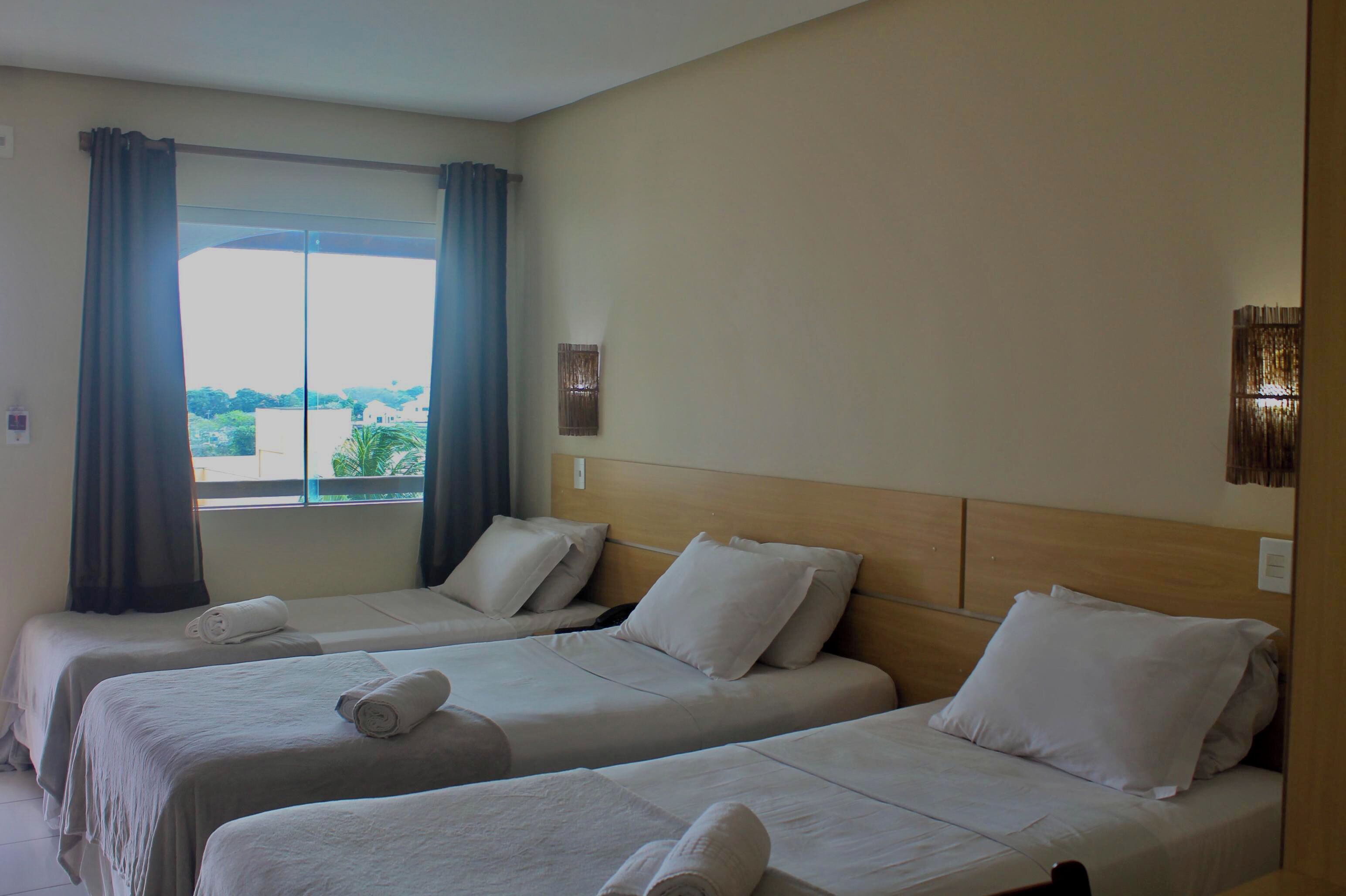 A hotel room with two beds, a large window with curtains, and a view of greenery outside.