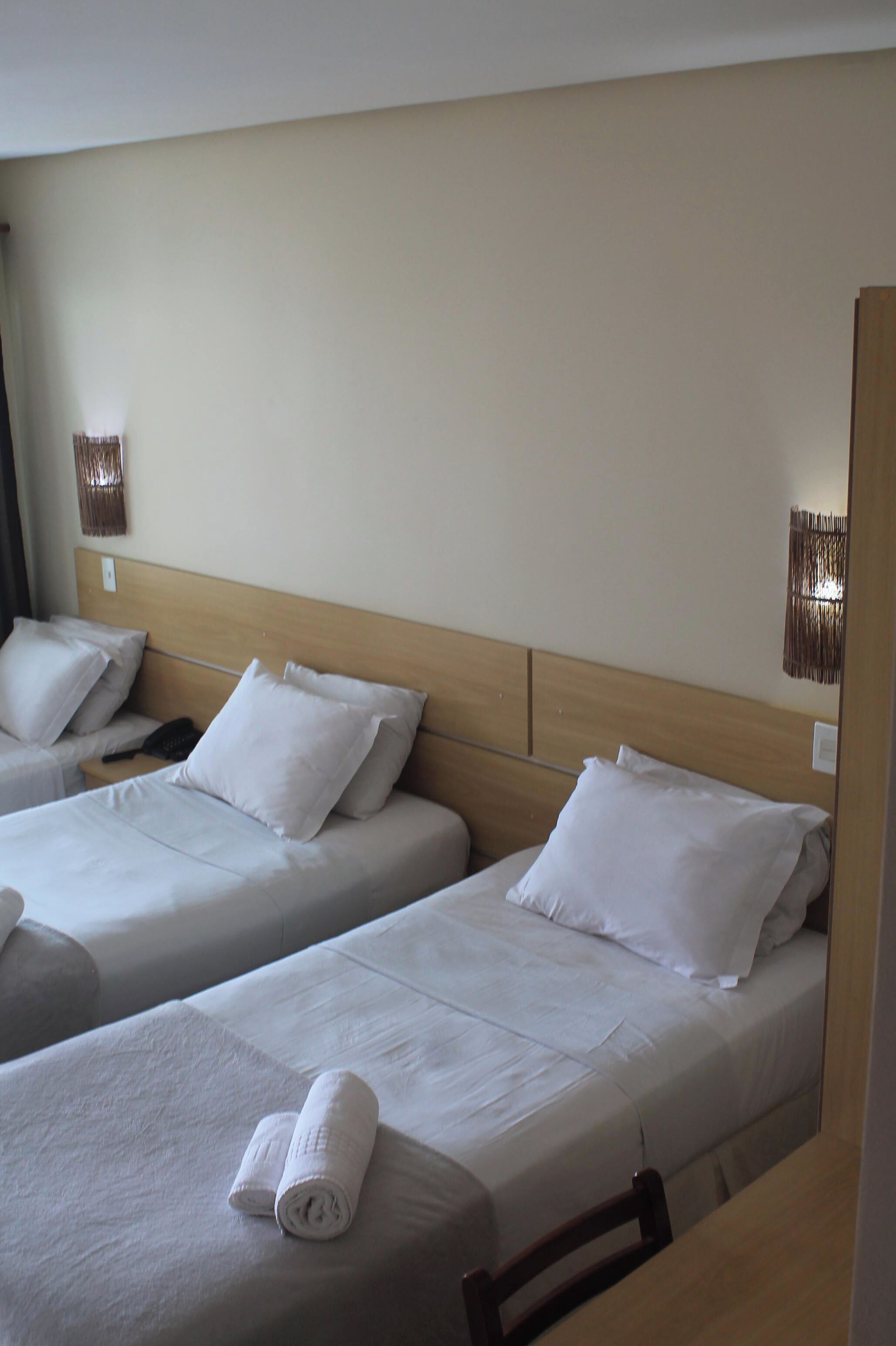 Two single beds with white linens and pillows, a wooden headboard, and a small table with a rolled towel.