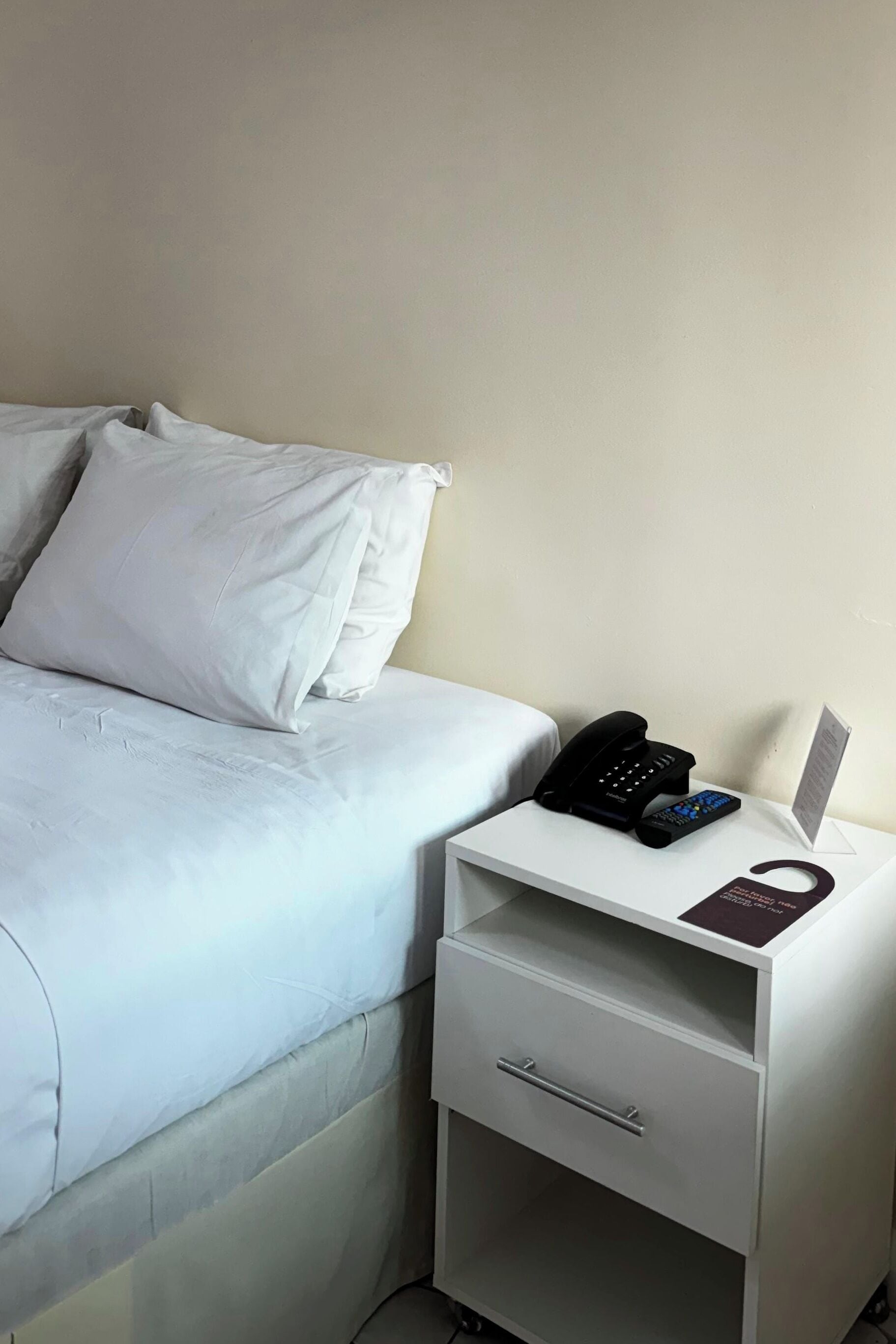 A hotel room with a bed, a white nightstand with a phone and remote control, and a small drawer.