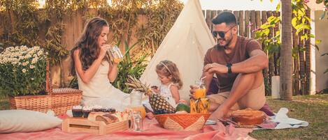 Un picnic familiar con una tienda de campaña, una cesta de picnic y comida extendida sobre una manta en un jardín trasero.