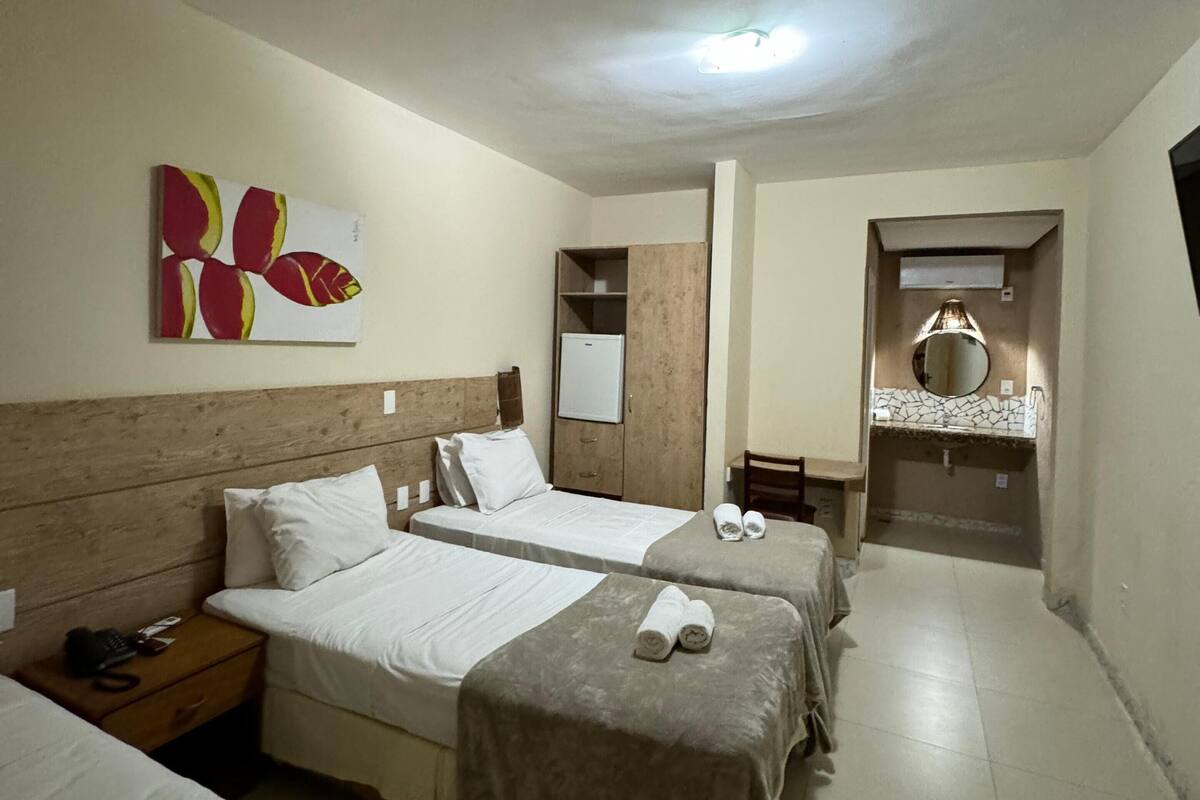A hotel room with two beds, a wooden headboard, a small bedside table with a phone, a wall-mounted mirror, and a bathroom visible through an open door.