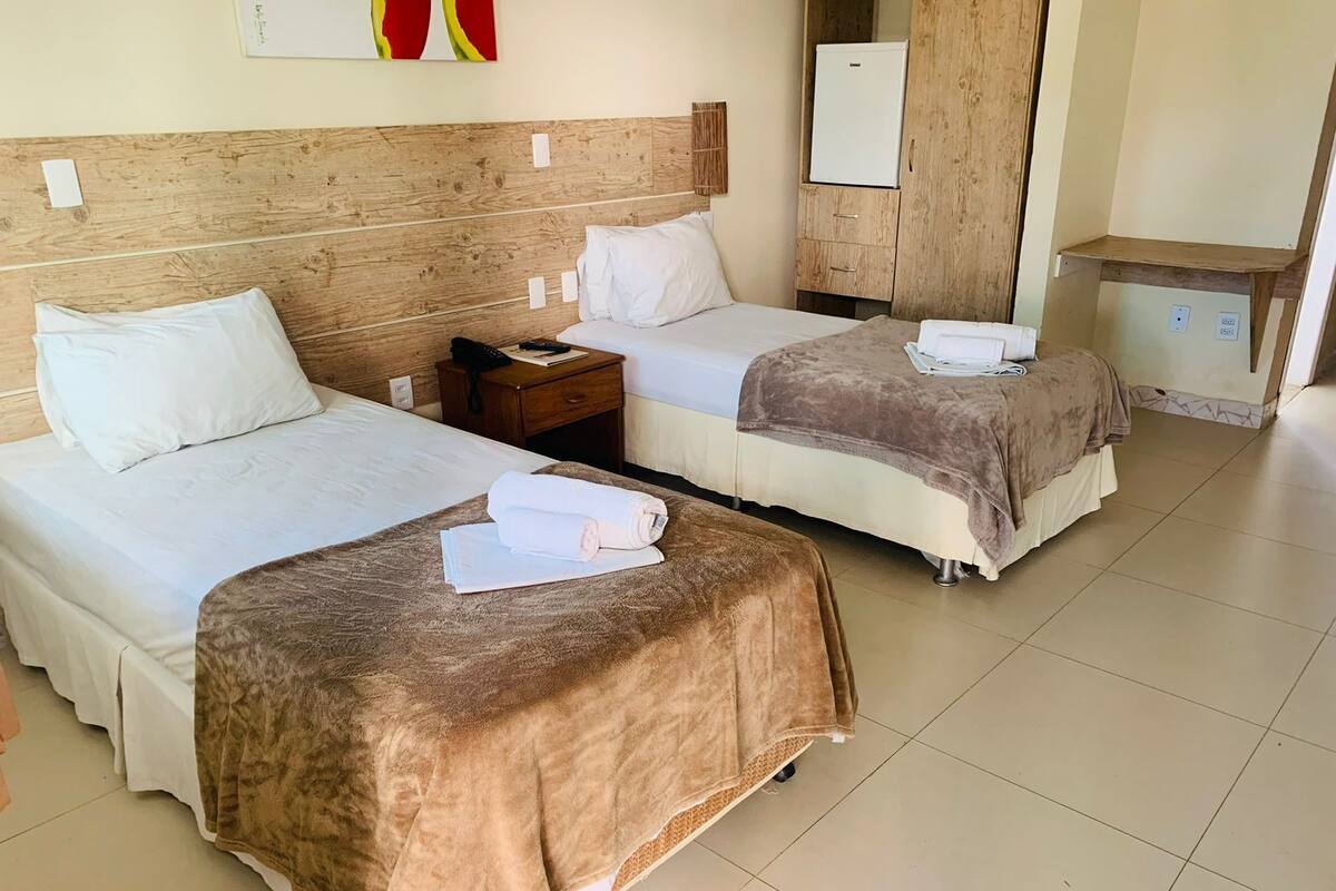 A hotel room with two beds, a wooden headboard, a small bedside table with a phone, a cabinet, and a refrigerator.