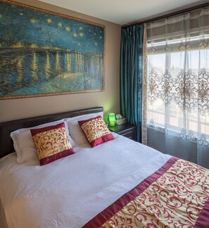 A hotel room with a large painting of a night scene, a bed with white linens and red and gold pillows, a nightstand with a green lamp, and a window with sheer curtains.