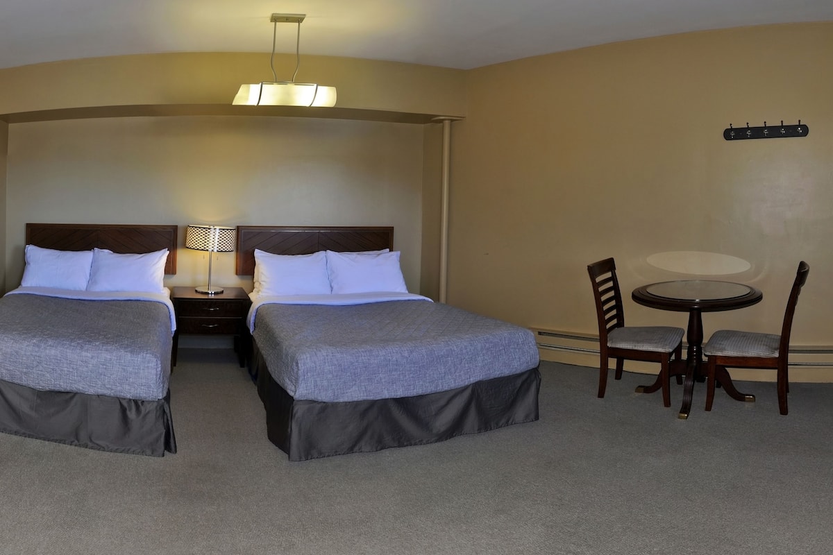 A hotel room with two beds, a nightstand, a table with chairs, a framed picture on the wall, and a ceiling light fixture.