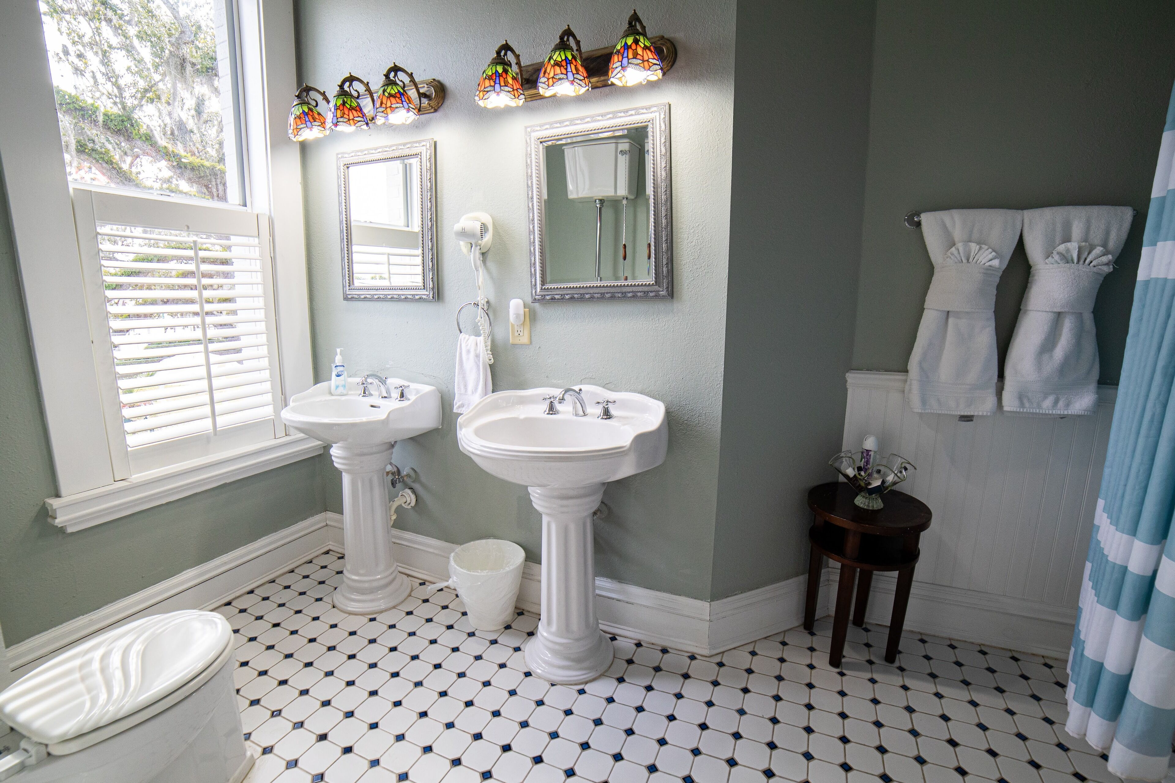 A bathroom with two sinks, a toilet, a window with blinds, a shower curtain, and a tiled floor.