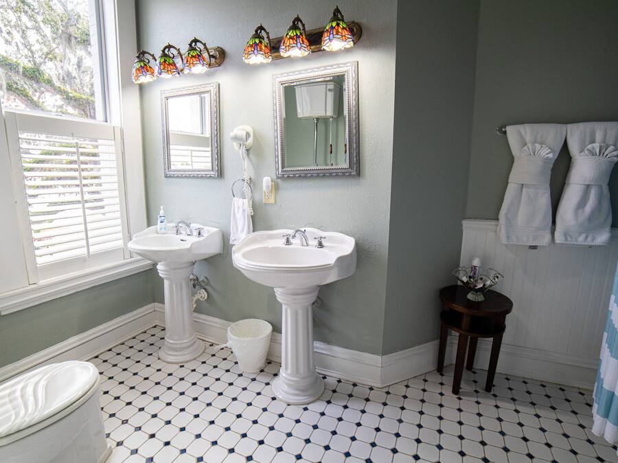 A bathroom with two sinks, a toilet, a window with blinds, a shower curtain, and a tiled floor.