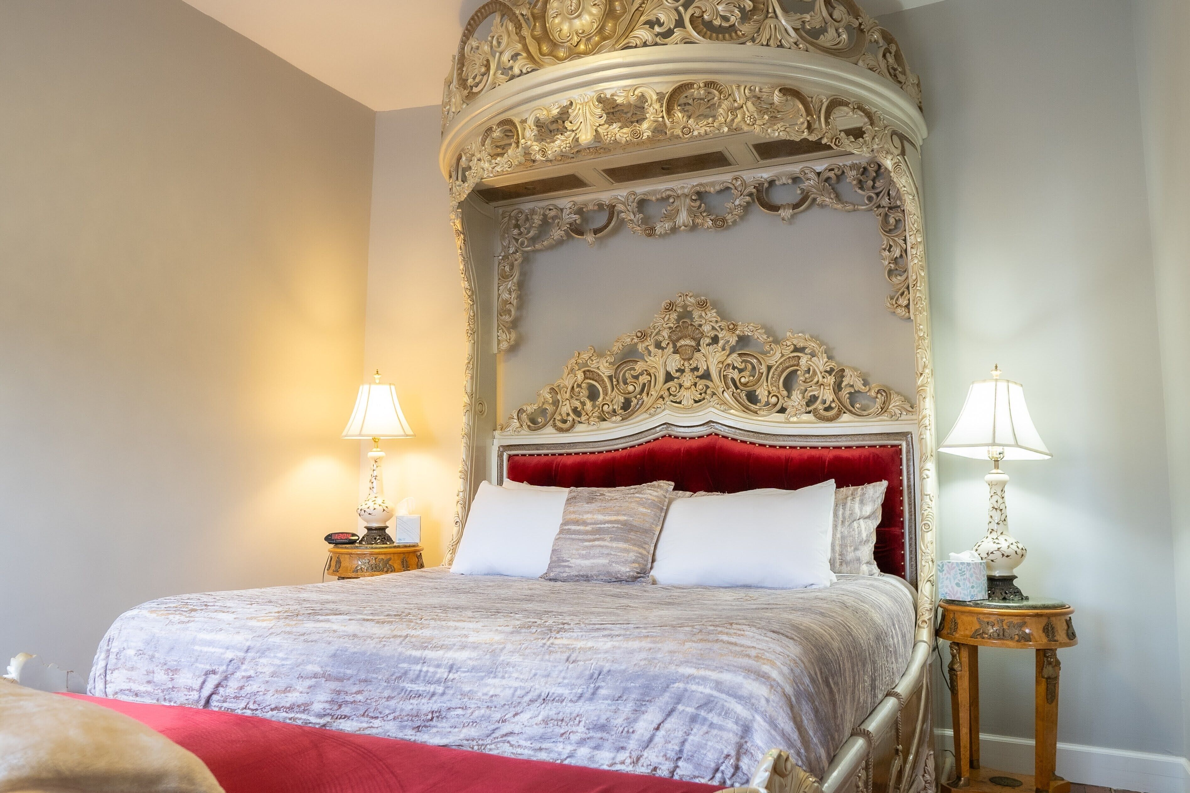 Uma cama com cabeceira vermelha e detalhes esculpidos, duas mesas de cabeceira com abajures, e um edredom estampado.