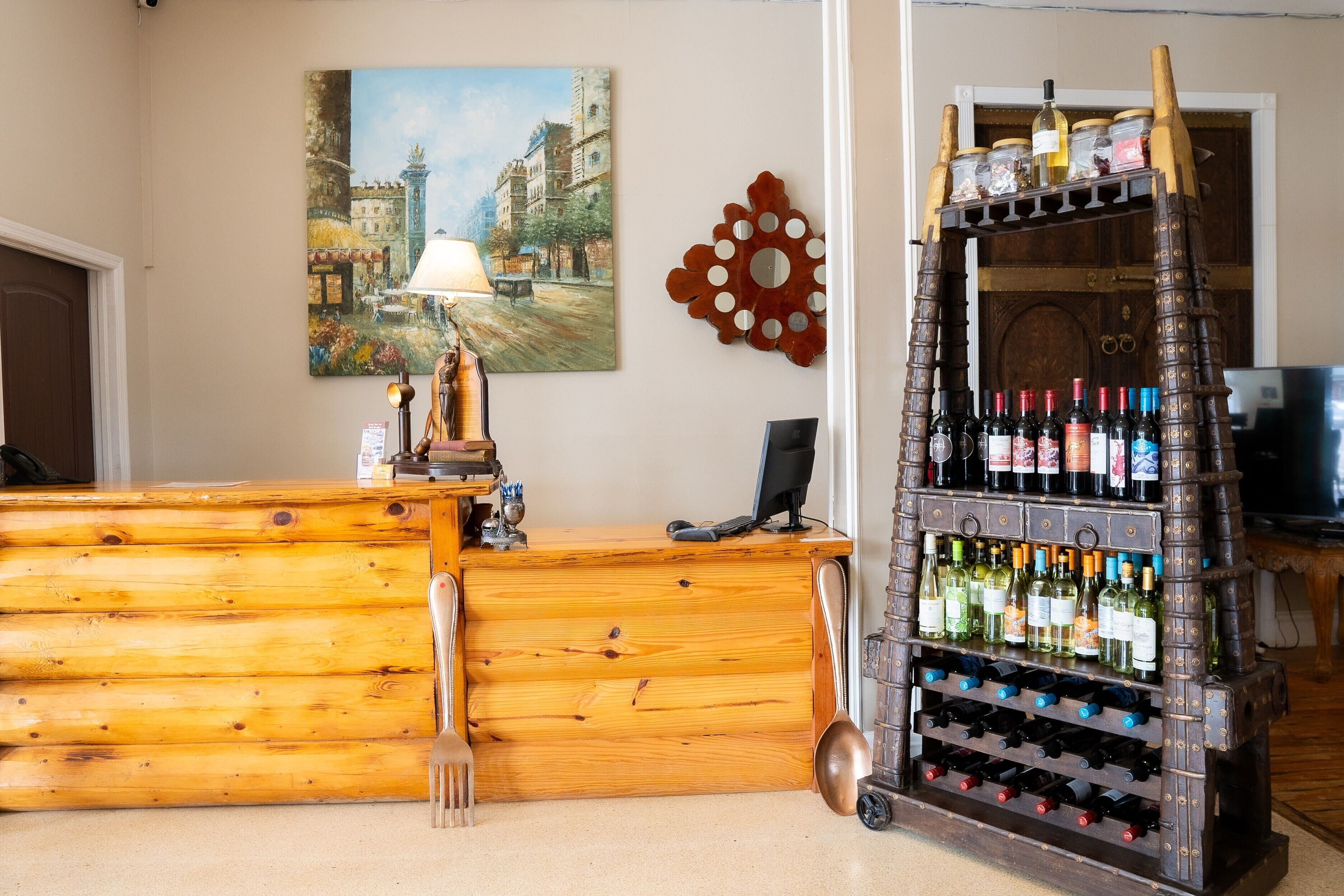 A wooden reception desk with a computer, a wine rack with bottles, a painting of a street scene, and a decorative mirror on the wall.