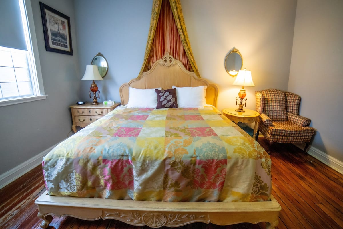 Ein Schlafzimmer mit einem großen Bett, einem Nachttisch, einem Stuhl, einem Spiegel und einem Fenster.