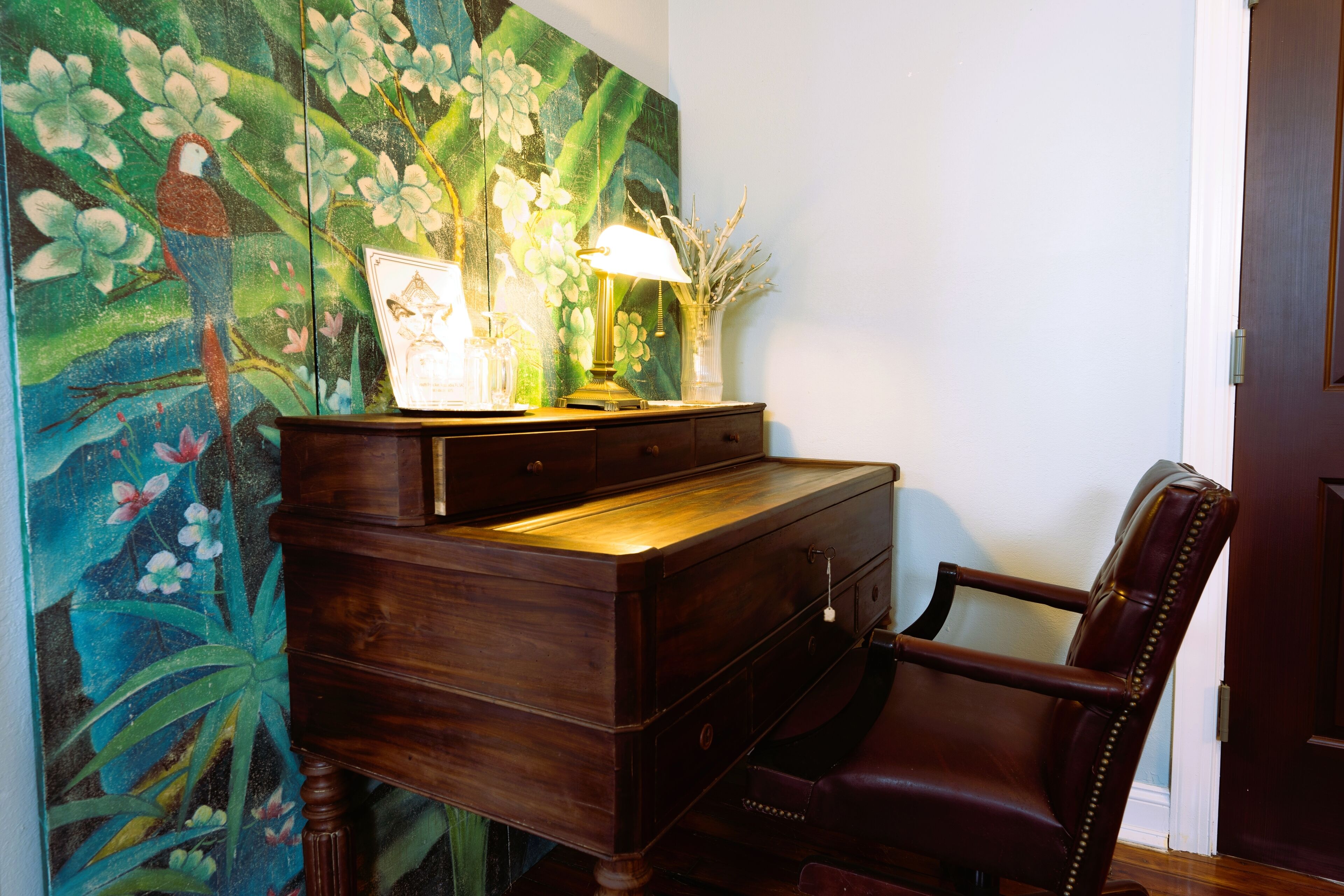 Un bureau en bois avec des tiroirs, une lampe et un tableau encadré. Une chaise en cuir est placée devant le bureau.