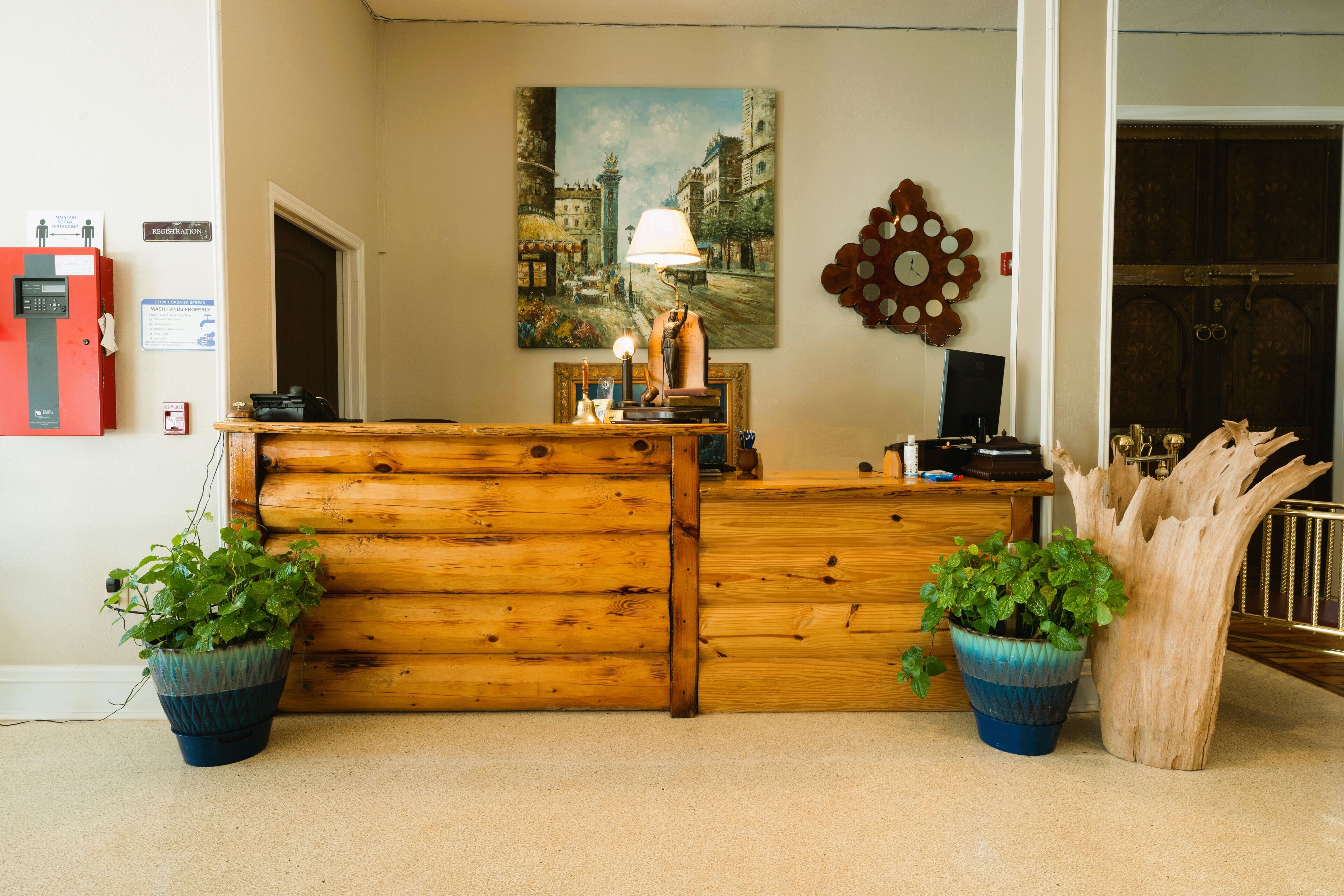 A wooden reception desk with a lamp, two potted plants, a painting of a cityscape, and a fire extinguisher.