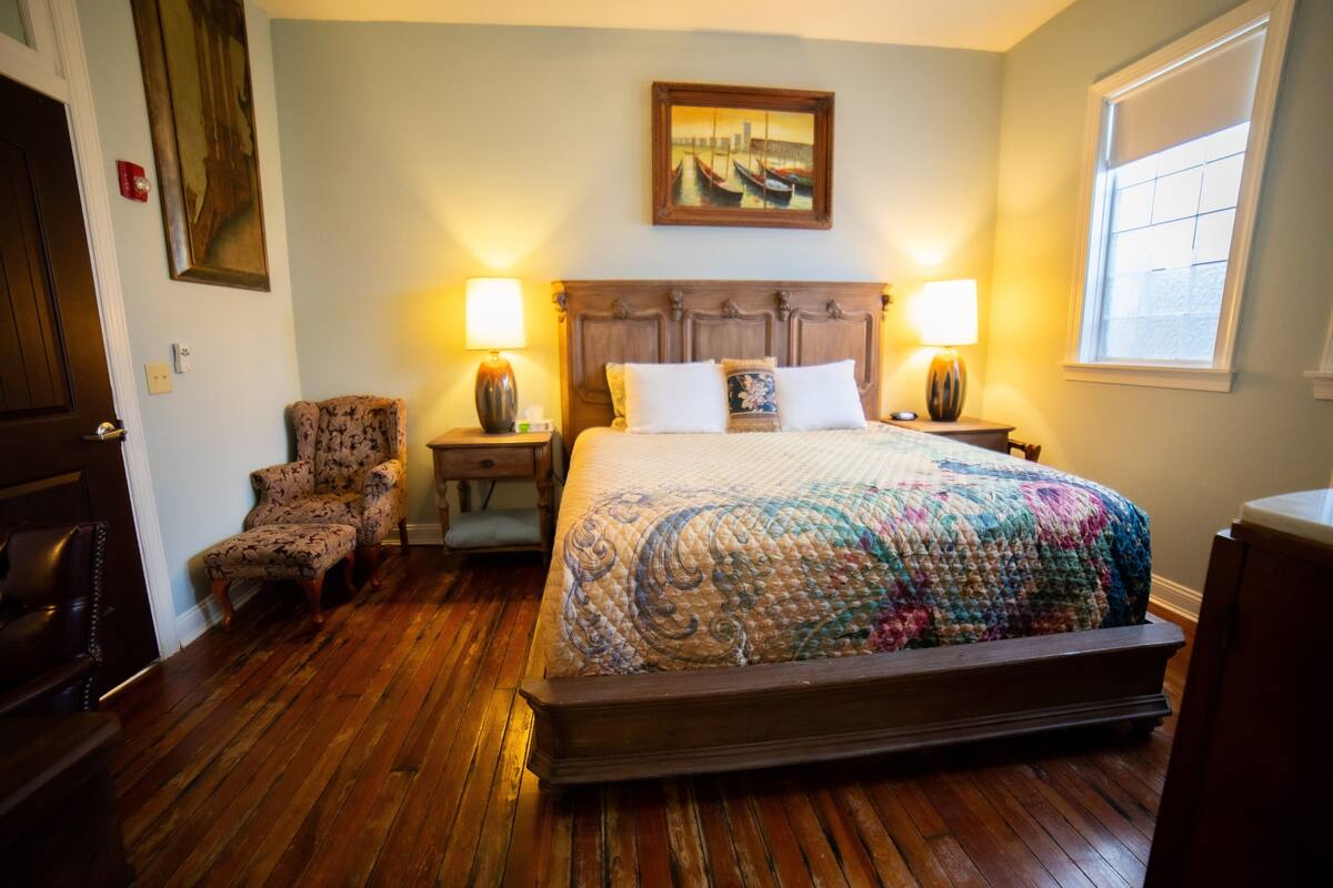 Ein Hotelzimmer mit Holzboden, einem großen Bett mit farbenfroher Steppdecke, zwei Nachttischlampen, einem Sessel und einem Bild an der Wand.